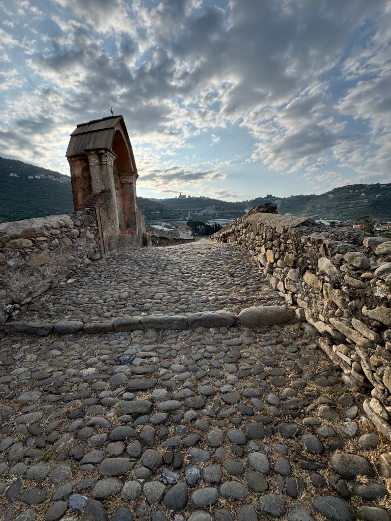 Ancient stone bridge in Taggia, Italy, featuring a cobblestone pathway and surrounding hills under a cloudy sky.